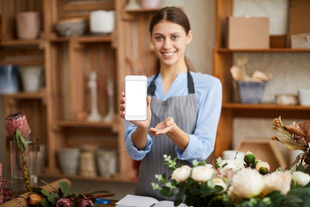 Businesswoman in a blue shirt and black apron holding a cell phone in a room with wooden shelves and white flowers