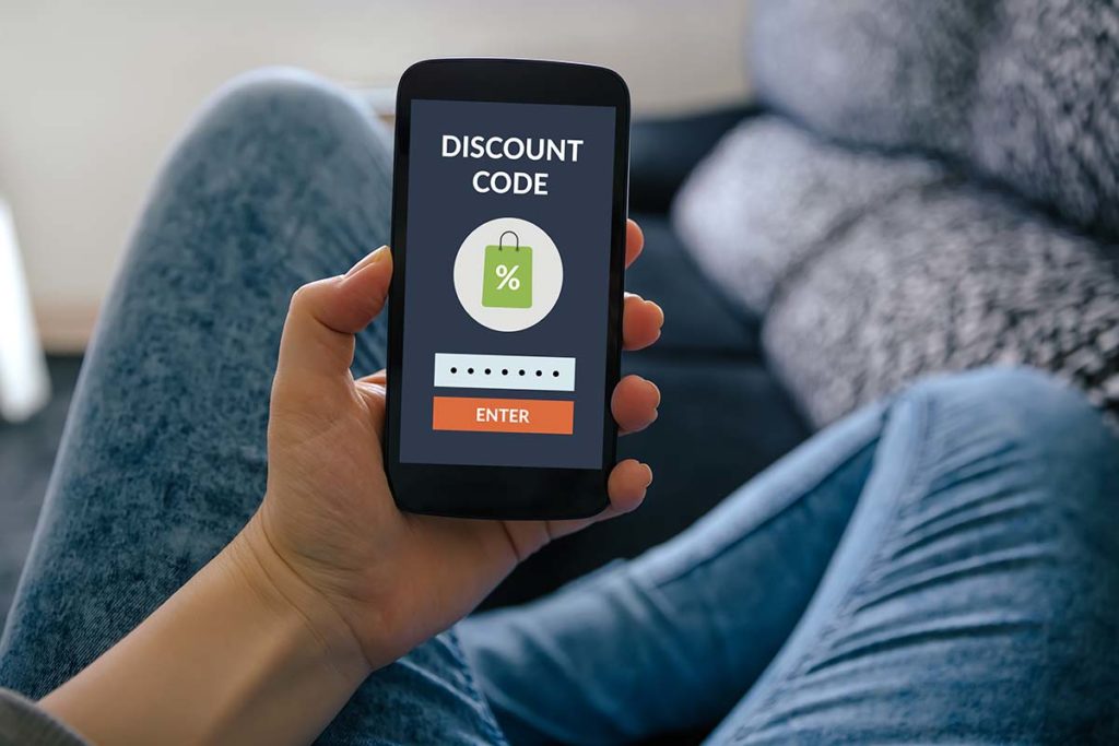 Person in blue jeans sitting cross-legged on a grey couch with a smartphone that has a discount code on the screen for shopping